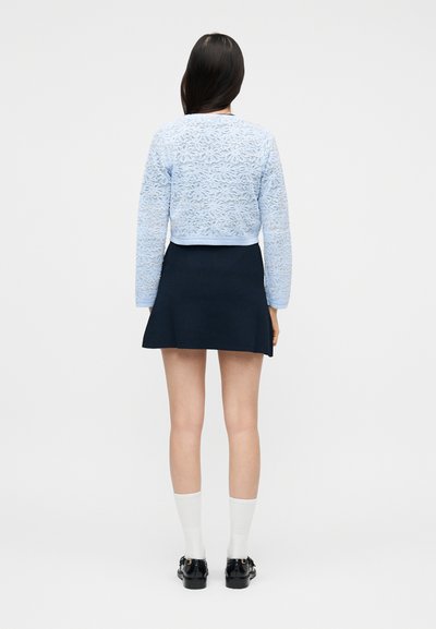 Γυναίκα που στέκεται και κοιτάζει προς τα πίσω, φορώντας μπλε φωτεινό δαντελωτό μακρυμάνικο τοπ, σκουρό μπλε φούστα σε σχήμα Α, λευκές κάλτσες και μαύρα μοκασίνια.