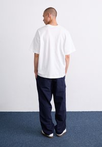 Hvid bomulds t-shirt med korte ærmer og en afslappet pasform, parret med marineblå cargobukser, vist fra bagsiden.