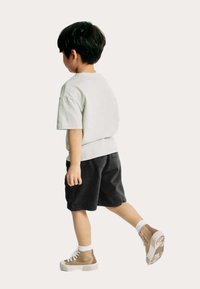 Un jeune portant un t-shirt gris clair et un short noir, avec des baskets montantes beiges. Le tissu semble doux ; le short a des poches sur les côtés.