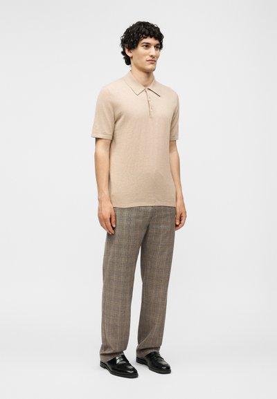 Tânăr stând, purtând un tricou polo bej cu mânecă scurtă, pantaloni maro în carouri și pantofi din piele negri pe un fundal gri deschis.