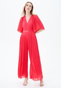 Mono rojo plisado con escote en V, mangas con vuelo y cintura ceñida con un cinturón que presenta un toque metálico. Tejido fluido y diseño de pierna ancha.