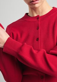 Cardigan en tricot rouge avec un col rond et cinq boutons foncés. Doté de poignets et d'un ourlet côtelés, mettant en valeur une texture lisse.