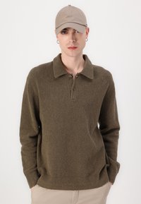 Brauner, strukturierter Pullover mit einem Kragen und zwei Knöpfen, kombiniert mit einer beigen Kappe. Der Stoff sieht weich und flauschig aus.
