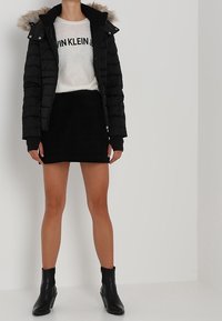 Czarna kurtka puchowa z kapturem wykończonym futrem, biała sweter z czarnym napisem, czarna zamszowa spódnica i czarne botki za kostkę. Widoczne gładkie tekstury.