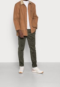 Brun kordflodjacka med en beige krage, vit t-shirt under, gröna kordflodbyxor och vita sneakers med grå och orange detaljer.