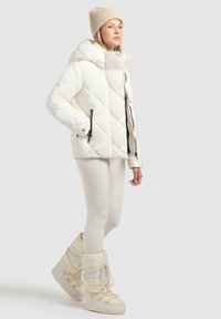 Witte gewatteerde pufferjack met een grote capuchon, voorzien van ritssluitingen in de zakken en een ontspannen pasvorm, gecombineerd met crèmekleurige high-top sneeuwlaarzen.