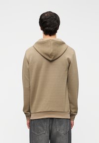 Μπεζ υφασμάτινο φούτερ με κουκούλα, διακοσμημένο με διαμάντι μοτίβο, ριμπ μανσέτες και τελείωμα. Συνδυάζεται με γκρι τζιν παντελόνι.