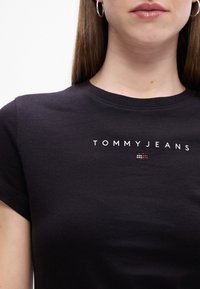 Melnas kokvilnas T-krekls ar īsām piedurknēm un apaļu kakla izgriezumu, kam raksturīgs balts "TOMMY JEANS" teksts un neliels sarkans, balts un zils logotipa elements.