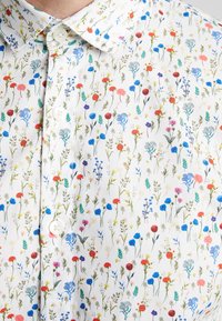 Camisa blanca de manga corta con un patrón floral que presenta flores rojas, azules y verdes, junto con un cuello clásico y cierres de botones.