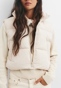 Kremasti prsluk s podstavom i zatvaranjem na patentni zatvarač, s uspravnim ovratom i prednjim džepovima, složen preko svijetlo bež strukturnog pulovera.