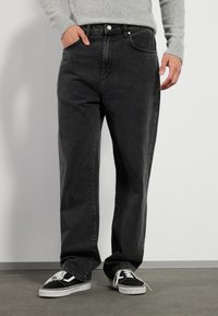 Svarte denimbukser med avslappet passform, med en tradisjonell femlommers design og synlig søm, kombinert med svarte sneakers.
