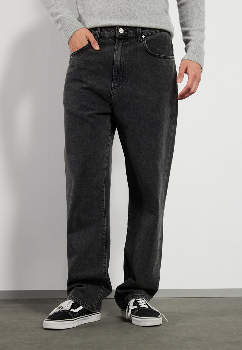 Svarte denimbukser med avslappet passform, med en tradisjonell femlommers design og synlig søm, kombinert med svarte sneakers.