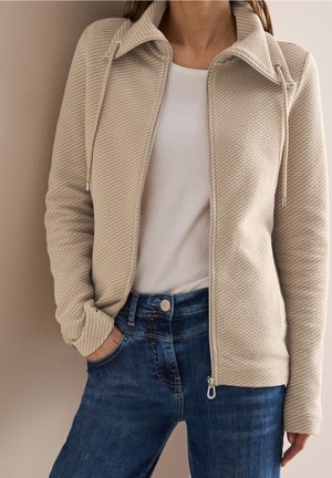 Frau trägt eine beige strukturierte Reißverschlussjacke über einem weißen Hemd und blauer Jeans, mit einer Hand in der Tasche vor hellem Hintergrund.