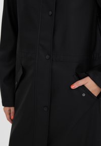 Black waterproof coat with snap buttons, two side pockets, and smooth matte texture. Features a sleek design and a minimalist aesthetic.