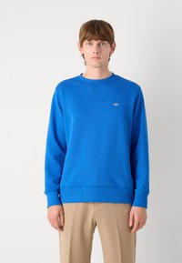 Blauw katoenen sweatshirt met een ronde hals en ribgebreide mouwuiteinden. Voorzien van een klein logo op de borst. Gedirigeerd met tan kleurige broeken.