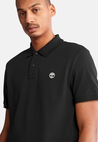 Black polo shirt made of textured cotton, featuring a ribbed collar, two buttons, and a white embroidered logo on the chest.