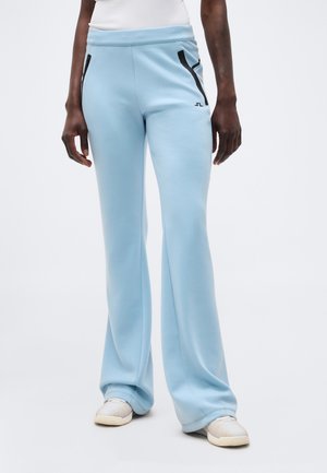 Partea inferioară a unei persoane care poartă pantaloni evazați de culoare albastru deschis cu detalii cu fermoar negru, top alb și adidași albi, stând pe un fundal alb.