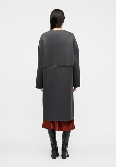 Γυναίκα με μακριά σκούρα μαλλιά που φοράει σκούρο γκρι μακρύ παλτό πάνω από φούστα σε χρώμα σκουριάς και μαύρες ψηλές μπότες έως το γόνατο, στέκεται με την πλάτη προς το μέρος μας σε λευκό φόντο.