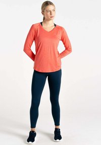 Langärmliges korallenfarbenes Sportshirt mit V-Ausschnitt, kombiniert mit navyfarbenen Leggings. Hat eine weiche Textur und eine figurbetonte Passform, getragen mit schwarzen Sneakern.