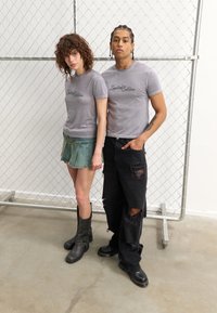 T-shirts gris avec "Édition Limitée" imprimé en écriture manuscrite, associés à une jupe en denim et un pantalon baggy noir déchiré ; des bottes robustes aux pieds.