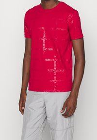 Rotes T-Shirt mit kurzen Ärmeln, das ein strukturiertes Muster des Wortes "ICEBERG" in Weiß aufweist. Rundhalsausschnitt, schmaler Schnitt, aus weichem Baumwollmaterial.