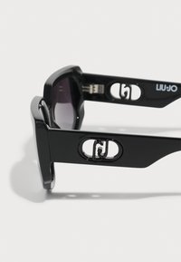 Gafas de sol negras con un acabado brillante, que presentan lentes de gran tamaño, detalles de logotipo recortados en las varillas y un diseño elegante y angular.