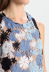 Spetsöverdel med blommönsterbroderi i nyanser av blått, svart och vitt, med en genomskinlig bas och rund halsringning. Exponerade sömnadsdetaljer.