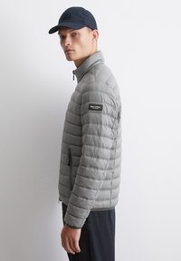 Grå pufferjakke med vandret quiltet tekstur, høj krave og sidelommer med lynlås. Båret med en marineblå kasket, minimal brandingdetalje på ærmet.