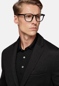 Black textured blazer over a black polo shirt with three buttons and a pocket. Clear glasses accentuate the design without additional patterns.