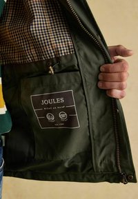 Ranka laiko atvirą žalią vandeniui nepralaidų �švarką, kurio vidinėje pamušalo pusėje matyti tarkuotas dizainas ir kišenė su Joules prekės ženklo etikete bei užtrauktuko detalėmis.