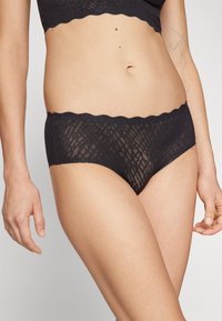 Culotte en dentelle noire avec des bords festonnés, présentant un motif texturé et une finition lisse. Conçue pour le confort et un ajustement près du corps.