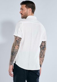 Weißes kurzärmliges Hemd aus strukturiertem Stoff, mit Kragen, seitlichen Schlitzen und Knöpfen an den Ärmeln. Sichtbare Tätowierungen am Arm.