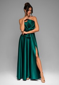 Abito maxi in raso verde smeraldo con design senza spalline, dettagli del corpetto annodati e uno spacco alto alla coscia. Completato da tacchi dorati.