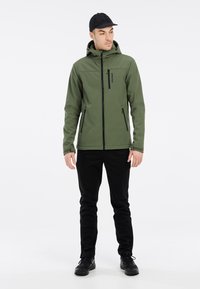 Grøn softshell-jakke med hætte, lynlåslukning foran og to sidelommer; parret med sorte bukser og sorte sko. Glat tekstur, strømlinet design.