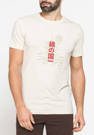 Uomo barbuto che indossa una t-shirt beige con caratteri giapponesi rossi e il contorno di un edificio a pagoda tradizionale.