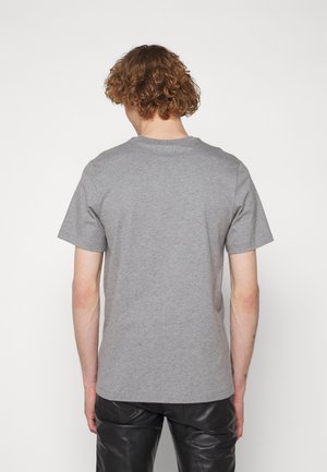 Personne aux cheveux roux bouclés portant un T-shirt gris uni à manches courtes et un pantalon en cuir noir, vue de dos sur un fond blanc.