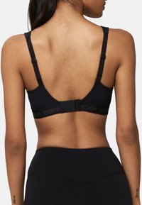 Brassière de sport noire avec panneaux en mesh, bretelles réglables et fermeture à agrafe. Tissu doux et bande avec logo sont des détails remarquables.