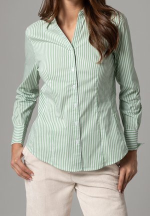 Kvinde iført en langærmet lysegrøn og hvid, vertikalt stribet skjorte med knapper og beige fløjlsbukser, hænderne hvilende på hoften.