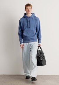 Giovane uomo in felpa blu e jeans chiari che cammina, portando una borsa da viaggio in pelle nera nella mano destra.