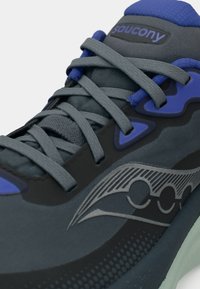 Ténis desportivos em cinza escuro com detalhes em azul, apresentando um material superior texturizado, atacadores planos e um padrão em forma de onda em cinza na lateral.