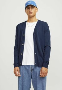 Cardigan blu navy con scollo a V, maniche lunghe e chiusura a bottoni, abbinato a una T-shirt bianca e jeans in denim, con una trama liscia.