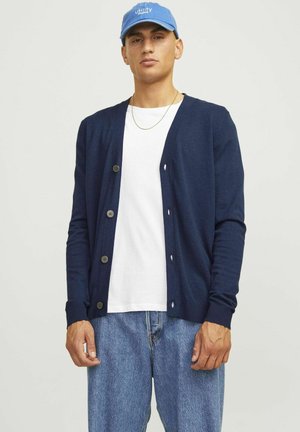 Cardigan blu navy con scollo a V, maniche lunghe e chiusura a bottoni, abbinato a una T-shirt bianca e jeans in denim, con una trama liscia.