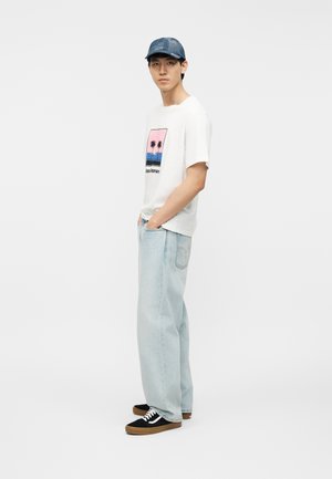 Νεαρός άνδρας με μπλε καπέλο, λευκό μπλουζάκι με εκτύπωση φοίνικα, ανοιχτό μπλε τζιν φαρδύ και μαύρα αθλητικά παπούτσια στέκεται με τα χέρια στις τσέπες.