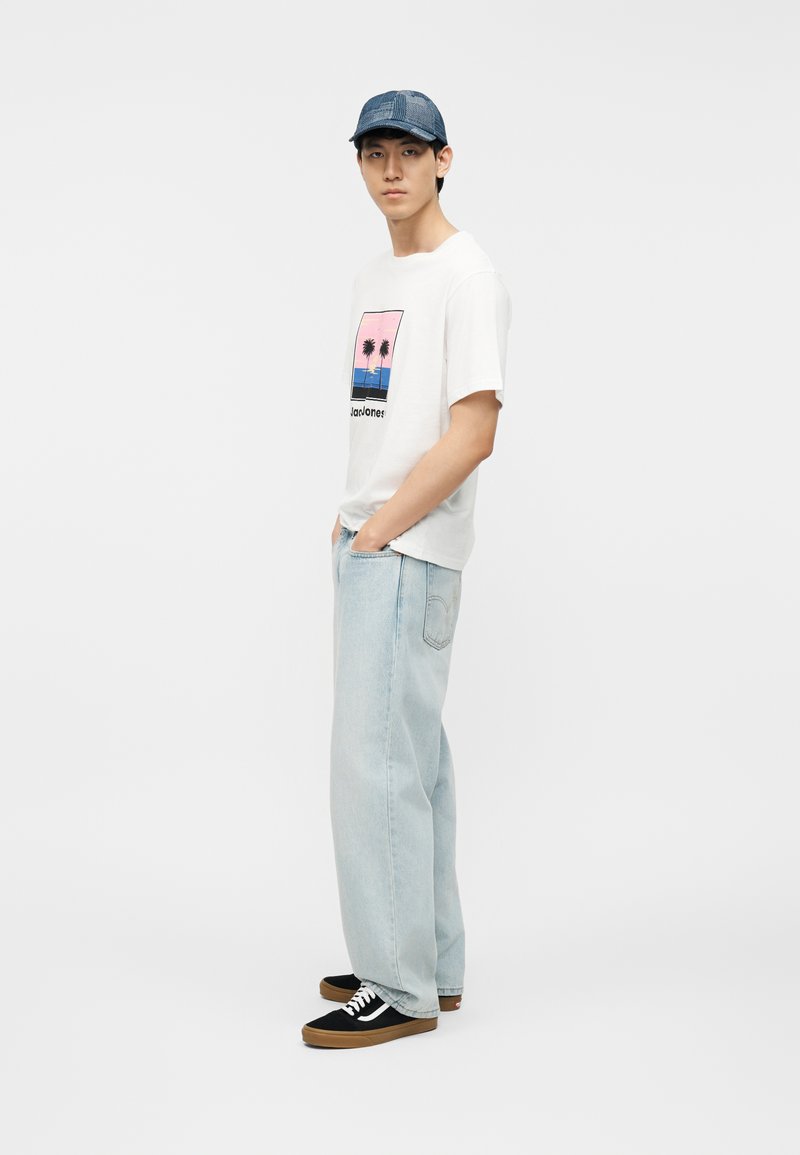 Young man in blue cap, white t-shirt with palm tree print, light blue wide-leg jeans, and black sneakers stands with hands in pockets.