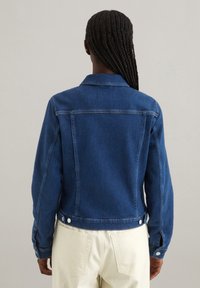 Blauwe spijkerjack met een getailleerd ontwerp, knoopmanchetten en een naaddetail op de achterkant. Beschikt over twee zijzakken en een verkorte lengte.