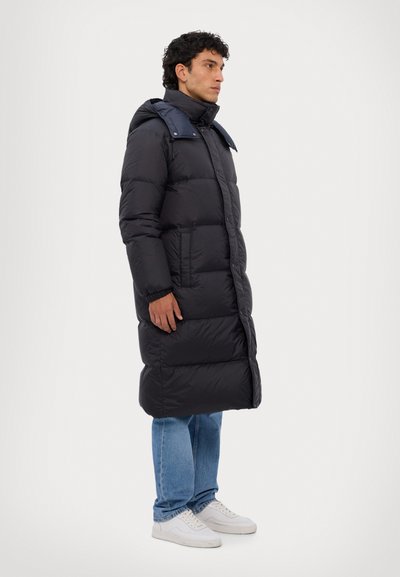 Lange zwarte pufferjas met een hoge kraag en blauwe accenten op de schouders; beschikt over twee zijzakken en ribbels bij de manchetten.