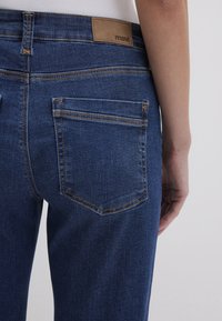 Dark blue denim jeans feature a tan leather label with "mavi" on the waistband, contrasting stitching, and two back pockets with rounded edges.