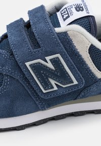 Baskets en velours bleu avec des accents en maille, arborant un logo "N" blanc, des bandes Velcro et une semelle en caoutchouc grise avec un motif de traction.