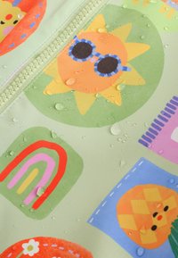 Mønstret stof med en lysegrøn baggrund, der har farverige illustrationer af en sol, regnbue og geometriske former. Vanddråber er synlige.