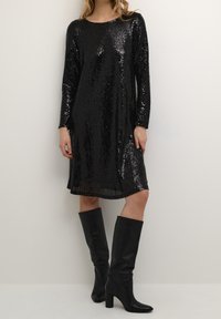 Kvinna som bär en svart långärmad paljettklänning och svarta knähöga klackstövlar, stående mot en enfärgad ljus vägg.